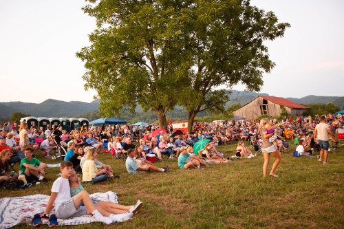 A large crowd gathers on a grassy field, sitting on blankets and enjoying an outdoor event with mountains in the background.
