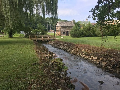 A serene landscape featuring a small stream, a wooden bridge, and a rustic cabin surrounded by greenery.