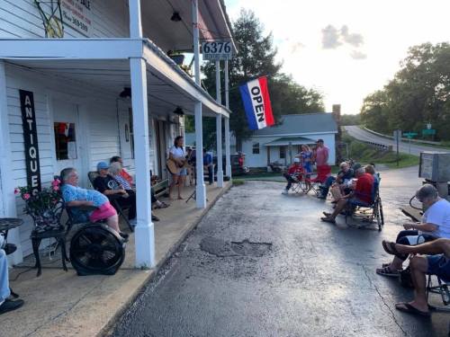 A group of people sitting outside an antique shop, enjoying live music on a warm evening.