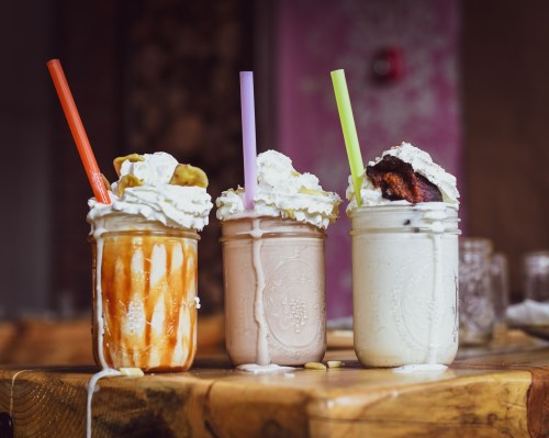 Three colorful milkshakes in jars topped with whipped cream, one with caramel, one chocolate, and one vanilla, on a wooden table.