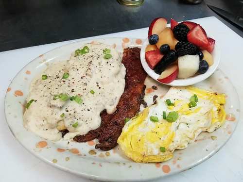 A plate of breakfast featuring biscuits and gravy, crispy bacon, a fried egg, and a side of mixed fruit.