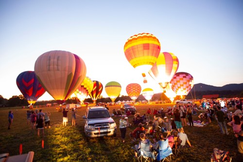 Colorful hot air balloons illuminate the sky at sunset, with spectators enjoying the scene on a grassy field.