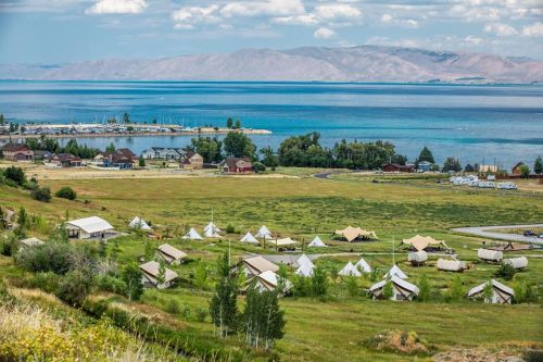 A scenic view of a lakeside area with tents, green fields, and mountains in the background under a partly cloudy sky.