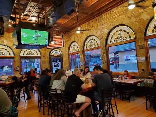 A bustling restaurant with patrons dining, large windows, and a TV showing a sports game. Warm, inviting atmosphere.