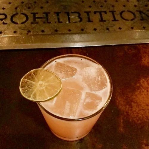 A cocktail with ice and a lime slice, placed on a bar with a "Prohibition" sign in the background.