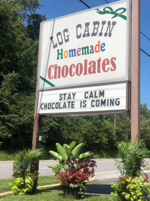 Sign for Log Cabin Homemade Chocolates with a message: "Stay Calm, Chocolate is Coming," surrounded by plants.
