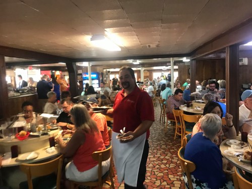 A busy restaurant scene with diners at tables and a waiter holding plates, creating a lively atmosphere.