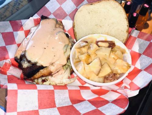 A sandwich with sauce on a checkered plate, accompanied by a side of potato salad in a cup.