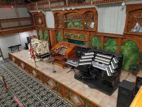A spacious stage featuring multiple organs and keyboards, surrounded by ornate wooden architecture and green decor.