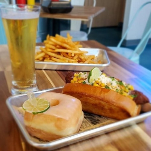 A tray with a donut, a sandwich topped with corn and lime, a side of fries, and a glass of beer on a wooden table.