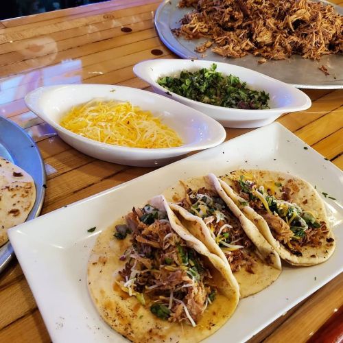 Three tacos filled with shredded meat, topped with cheese and herbs, served alongside bowls of cheese and salsa.