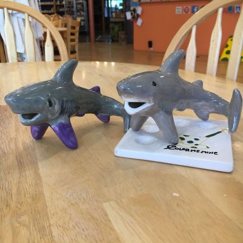 Two ceramic shark figurines, one with purple fins, sitting on a white base with a green design, on a wooden table.