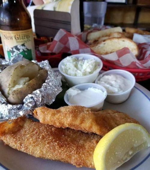 A plate with fried fish, a baked potato, coleslaw, and dipping sauces, with a beer and bread in the background.