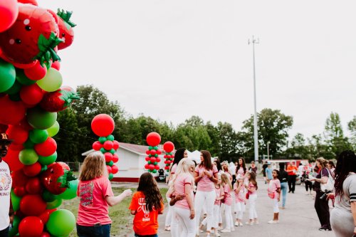 A festive scene with people in pink shirts and white pants, celebrating under colorful balloon arches.