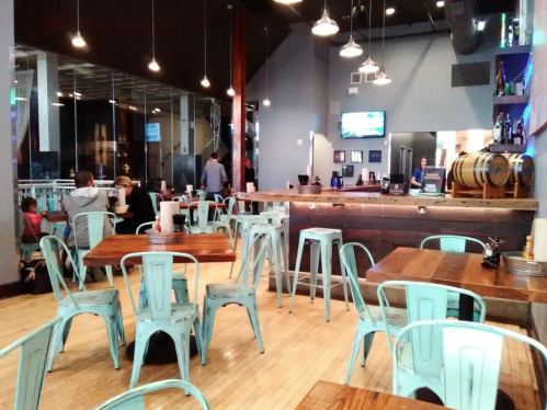 A modern restaurant interior with wooden tables, metal chairs, and a bar area, featuring a casual atmosphere.