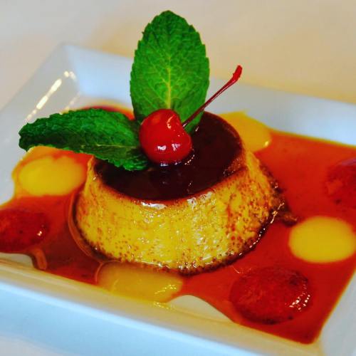 A creamy dessert topped with a cherry and mint leaves, surrounded by colorful fruit sauces on a white plate.