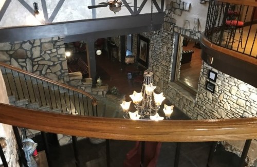 A view from above of a spacious, rustic interior with stone walls, a chandelier, and a curved wooden railing.