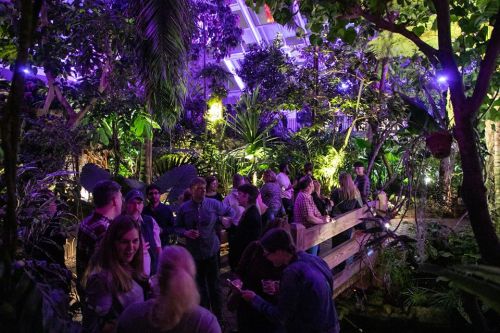 A vibrant indoor garden party with guests mingling among lush plants and colorful lighting.