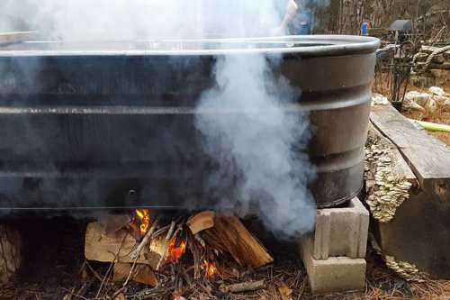 A metal tub sits on blocks, with a fire burning underneath, producing smoke in a wooded outdoor setting.