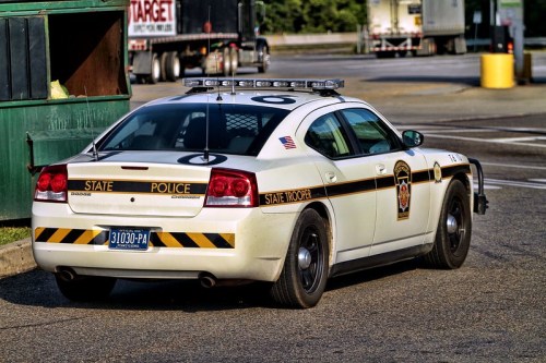 A white state police car with a black stripe parked near a dumpster in a parking lot.