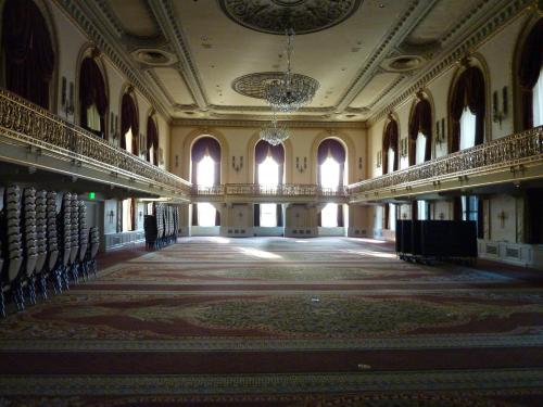 A spacious, elegantly decorated ballroom with chandeliers, large windows, and stacked chairs along the walls.