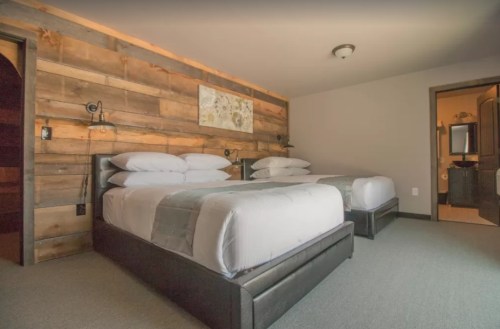 A cozy hotel room featuring two beds, wooden accent walls, and a bathroom door in the background.