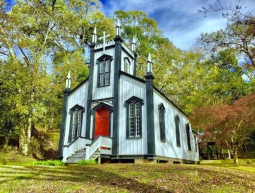 A quaint, Gothic-style church with a red door, surrounded by lush greenery and trees under a blue sky.