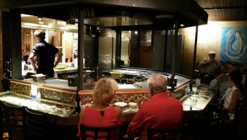 A couple sits at a restaurant counter watching a chef cook at a teppanyaki grill, with other diners in the background.