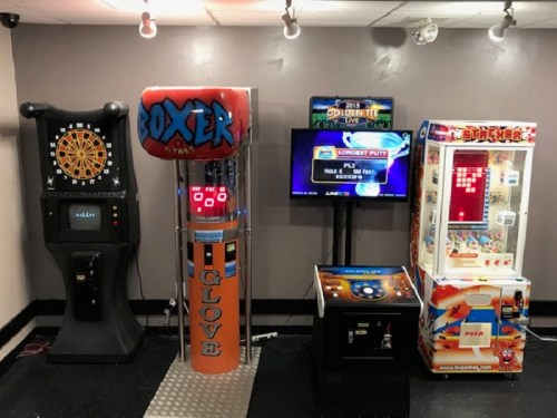 A game room featuring a dartboard, boxing machine, and various arcade games with colorful screens and lights.