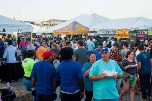 A crowded outdoor event with people mingling under tents, enjoying food and drinks in a lively atmosphere.
