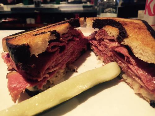 Close-up of a stacked sandwich with layers of meat, served with a pickle on the side. Toasted bread with a crispy edge.