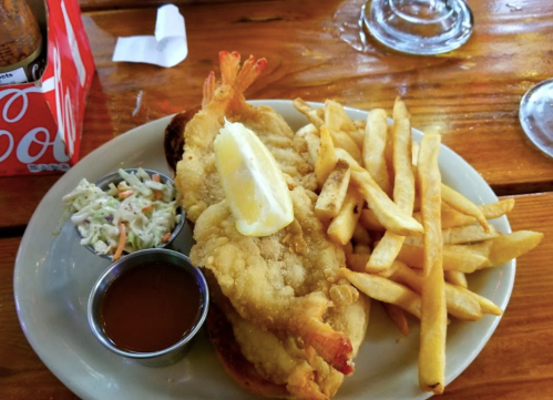 A plate with fried shrimp topped with lemon, coleslaw, fries, and a small cup of dipping sauce on a wooden table.