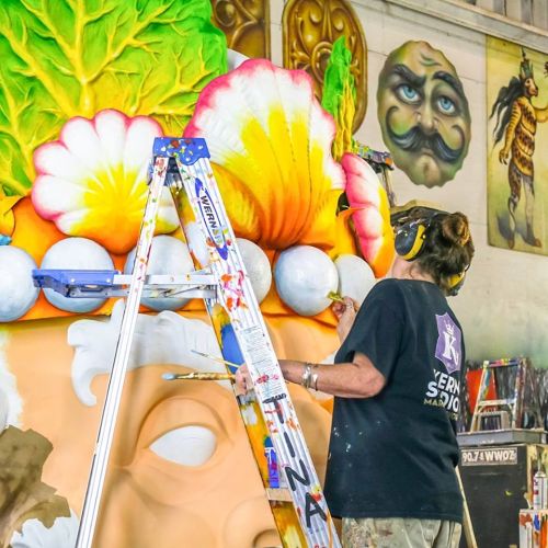 An artist paints a colorful mural on a wall, using a ladder and wearing headphones for protection.