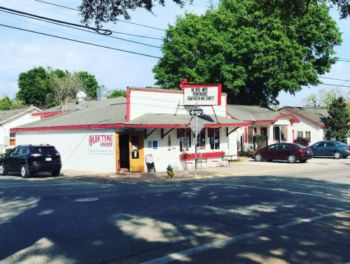 A quaint building with red trim, labeled "Quick Time Grocery," situated at a street corner with parked cars and trees nearby.