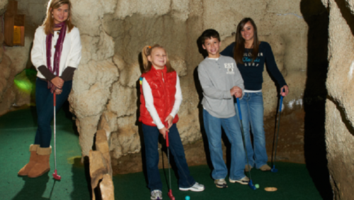 Four children are playing mini-golf in a cave-like setting, holding putters and smiling.