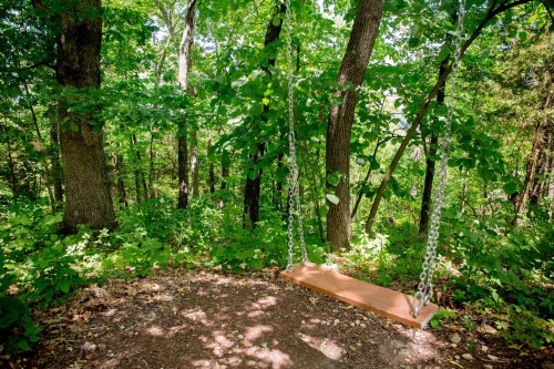 A wooden swing hangs from chains in a lush green forest, surrounded by trees and foliage.