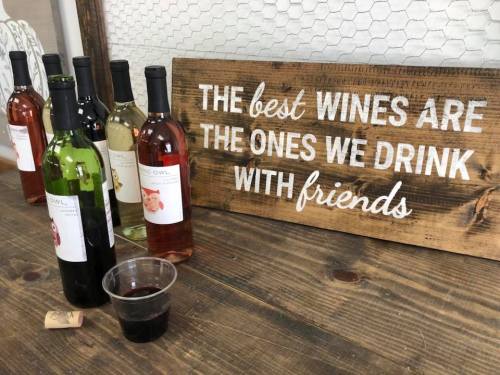 A wooden sign reads, "The best wines are the ones we drink with friends," next to several bottles of wine and a cup.