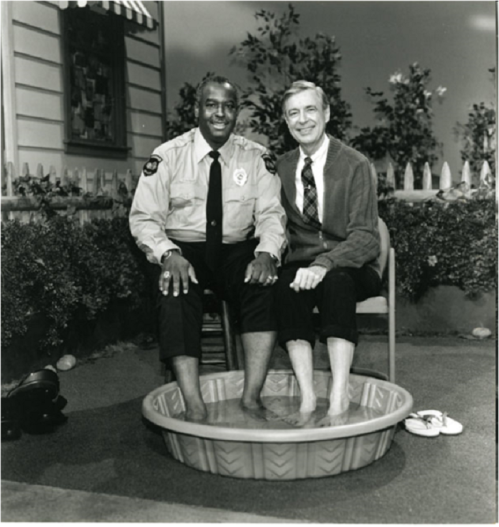 Black and white photo of two men sitting together in a foot bath, smiling, surrounded by greenery.