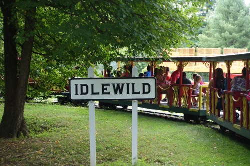 Sign for Idlewild amusement park with a train and people in the background, surrounded by trees.