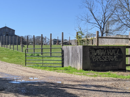 A rustic entrance to Barn Hill Preserve, featuring a wooden gate and a grassy field under a clear blue sky.