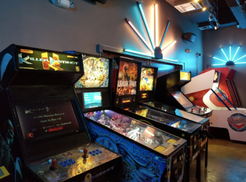A row of vintage arcade machines and pinball games illuminated by colorful neon lights.