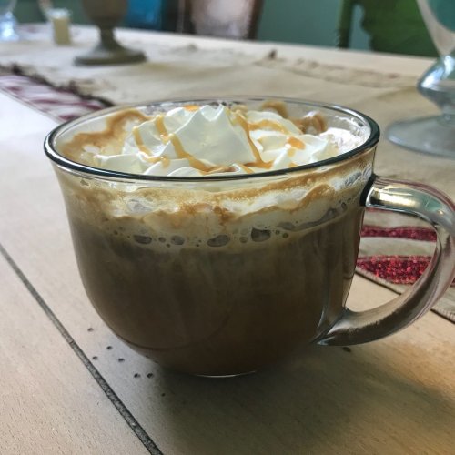 A close-up of a glass mug filled with coffee topped with whipped cream and caramel drizzle, set on a wooden table.