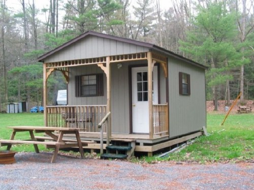 A small, rustic cabin with a porch, surrounded by trees and a grassy area, featuring picnic tables in front.
