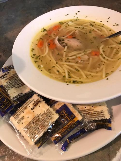 A bowl of chicken noodle soup with carrots and herbs, accompanied by packets of crackers on a plate.