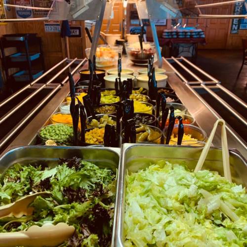 A vibrant salad bar featuring various toppings, including greens, olives, cheese, and dressings, in a rustic setting.