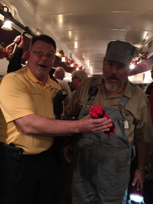 Two men in a train car, one in a yellow shirt and the other in overalls, holding a toy dynamite stick, looking surprised.