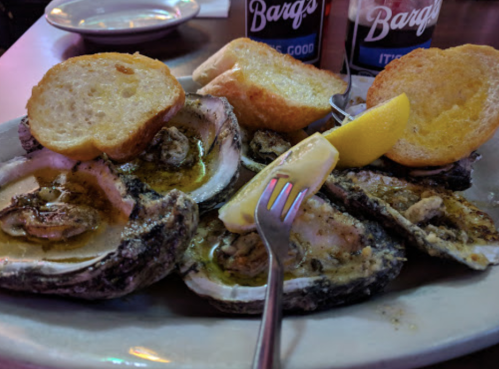 A plate of grilled oysters topped with garlic butter, served with lemon wedges and slices of toasted bread.