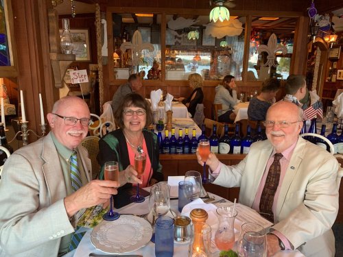 Three people toast with drinks at a restaurant, surrounded by tables and decor, smiling and enjoying their meal.