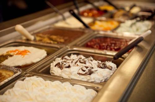 A dessert bar featuring various toppings and whipped cream in metal trays, with spoons for serving.
