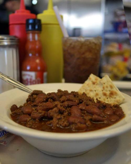 A bowl of chili with beans, served with crackers, and condiments in the background, alongside a drink.